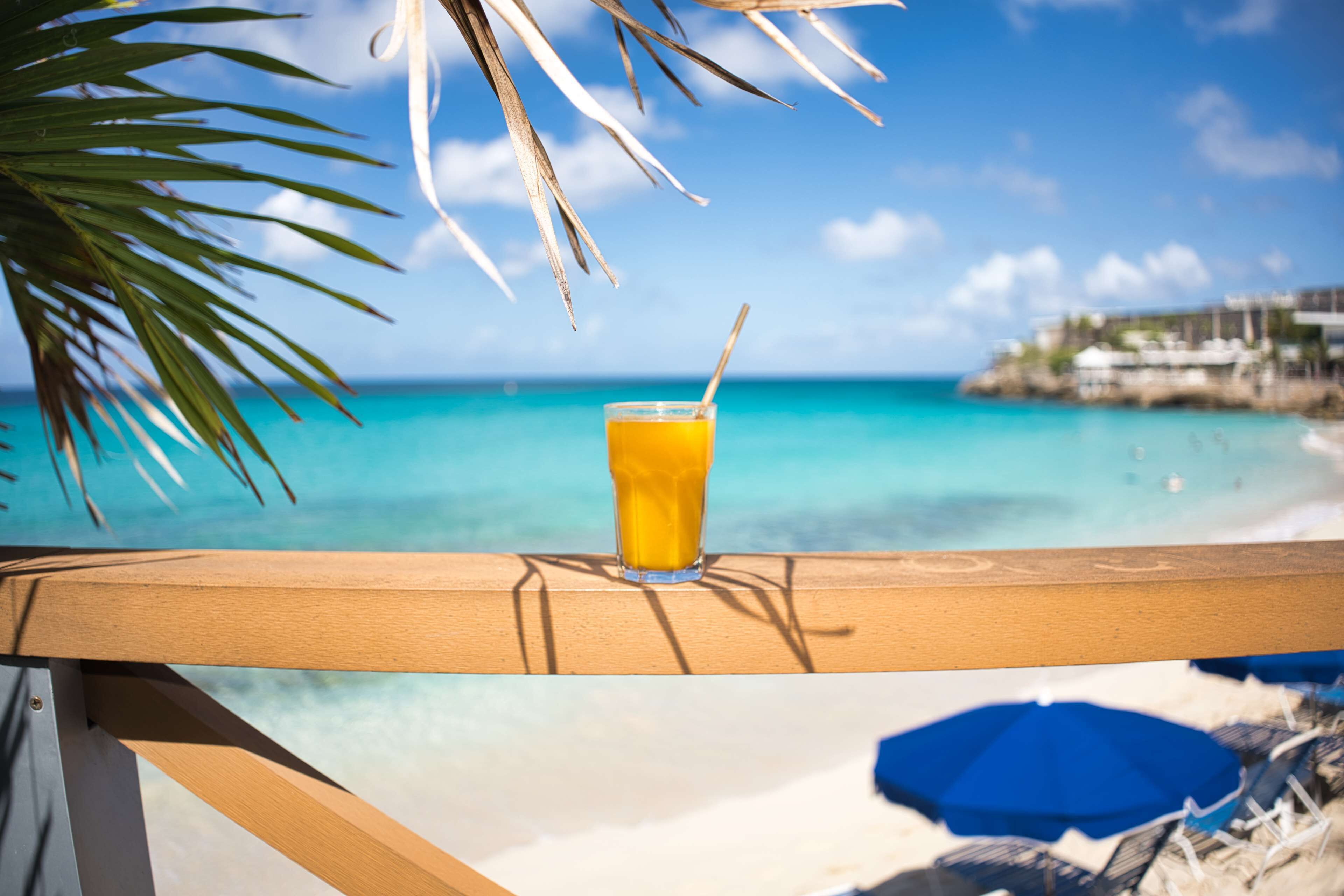 A glass of orange juice on a wooden railing with a view of a beach and ocean.