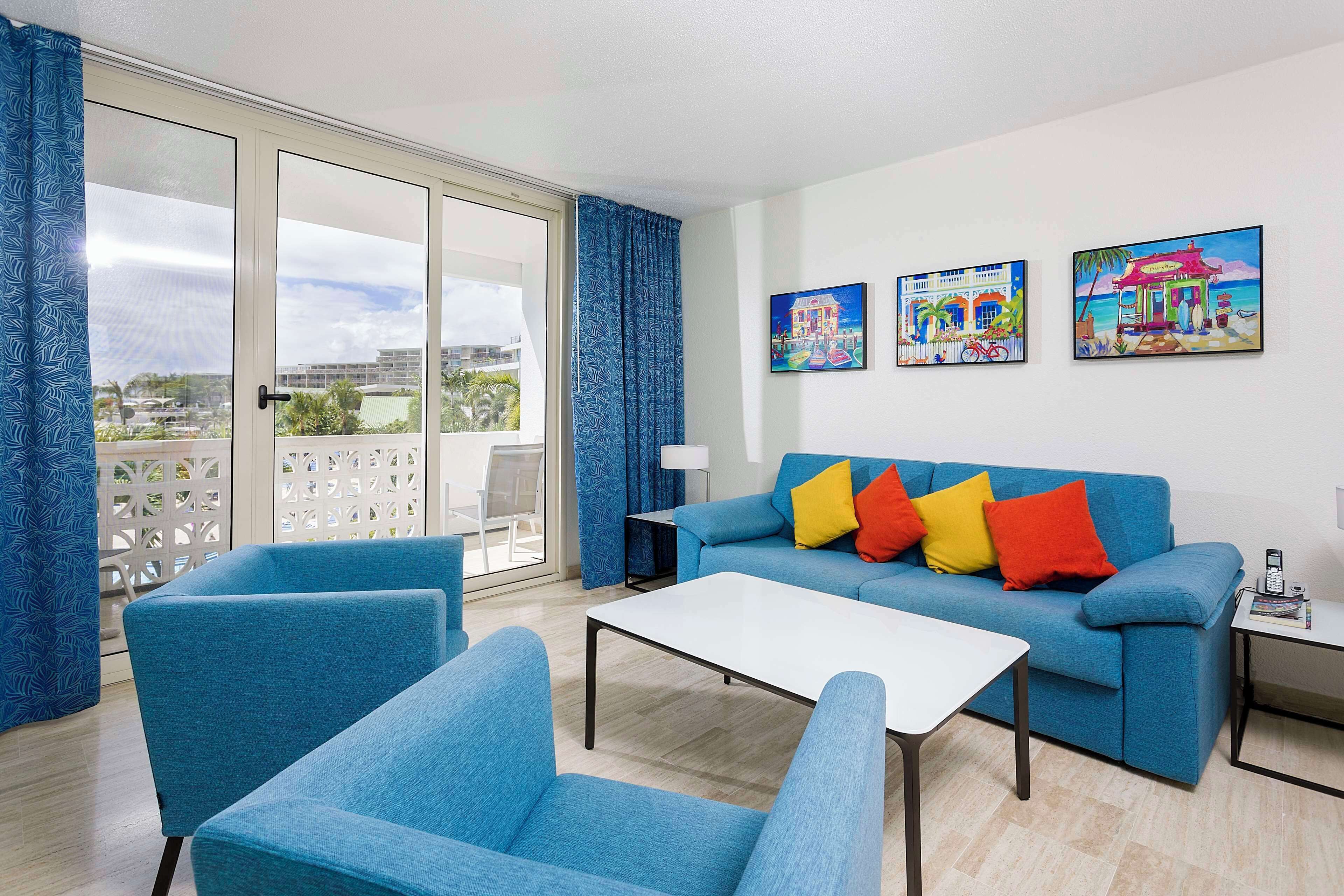 Una sala de estar con un sofá azul, sillones a juego, una mesa de centro blanca y un balcón con cortinas azules.