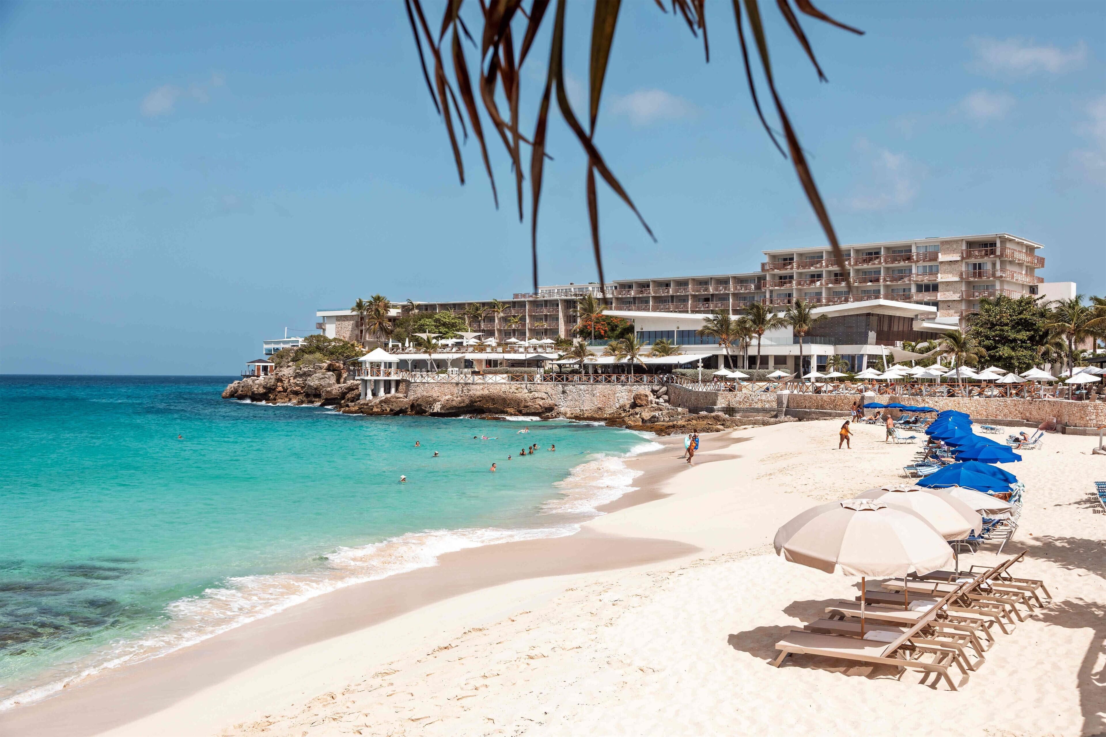A beach with clear turquoise water, a sandy shore, and a hotel with multiple balconies overlooking the sea.