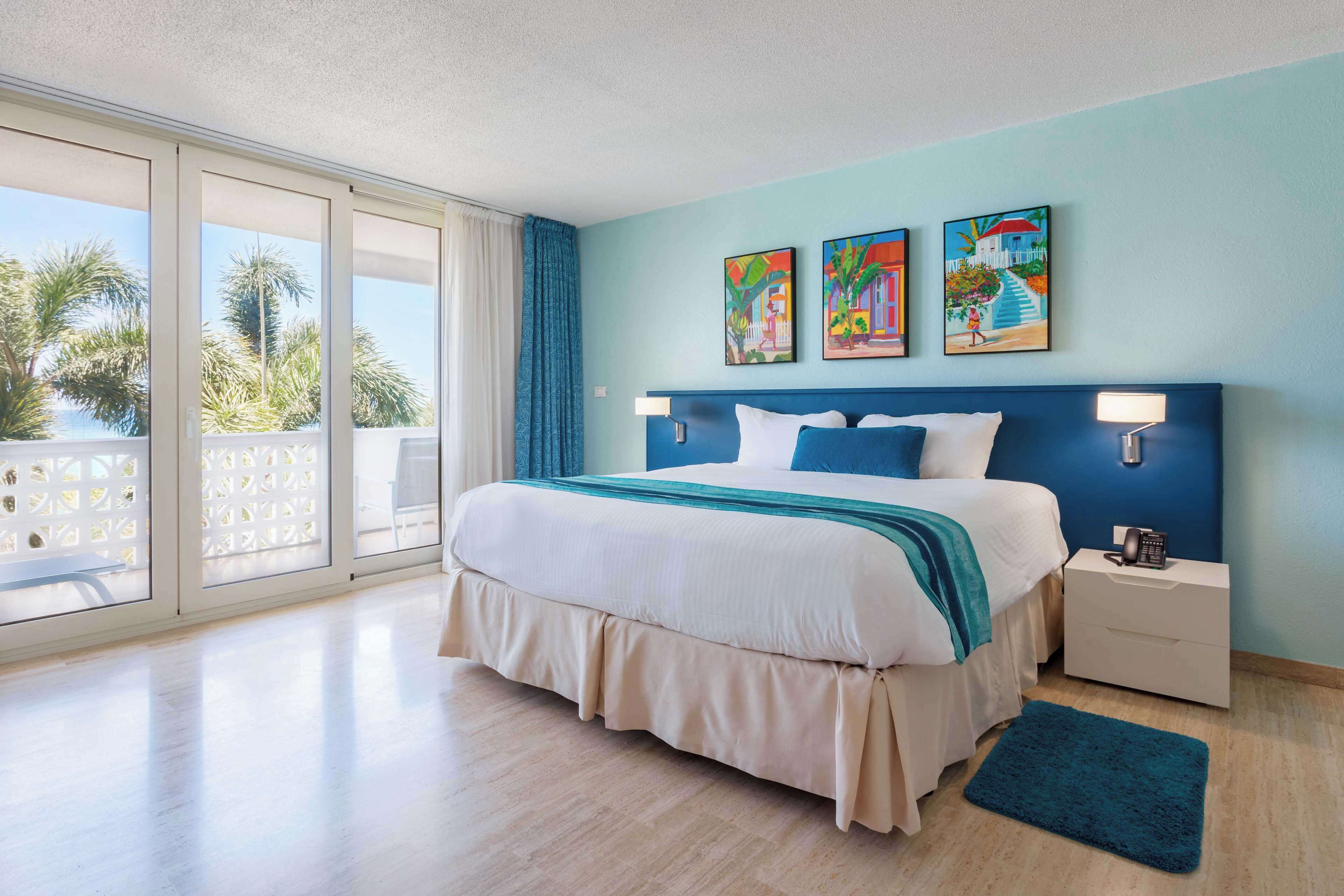A hotel room with a large bed, a nightstand, a blue rug, and a balcony with a view of palm trees.