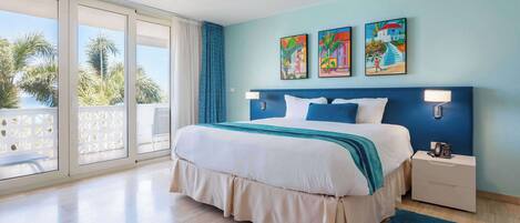 A hotel room with a large bed, a nightstand, a blue rug, and a balcony with a view of palm trees.