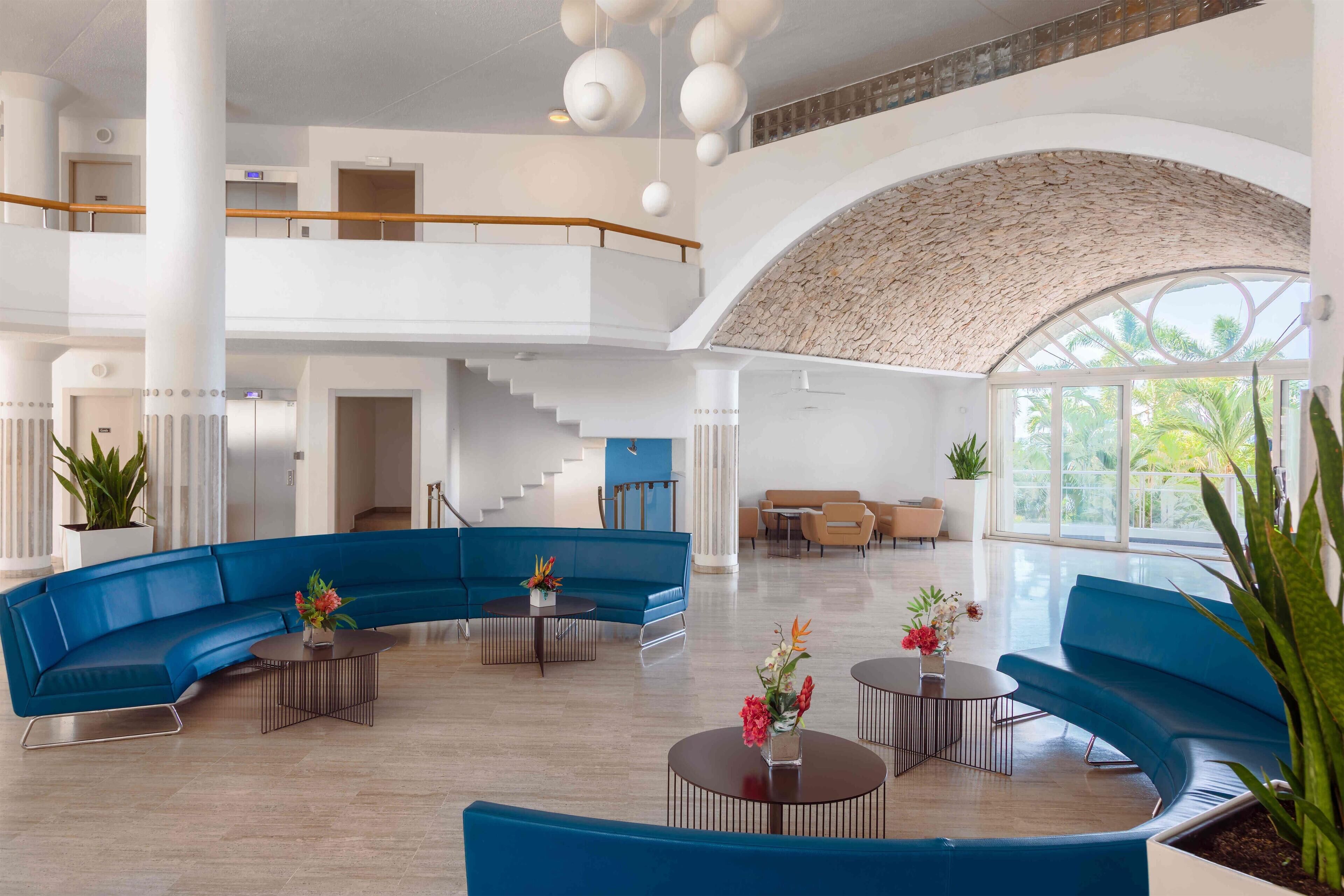 A spacious lobby with a curved blue sofa, round tables, and a staircase leading to an upper level.