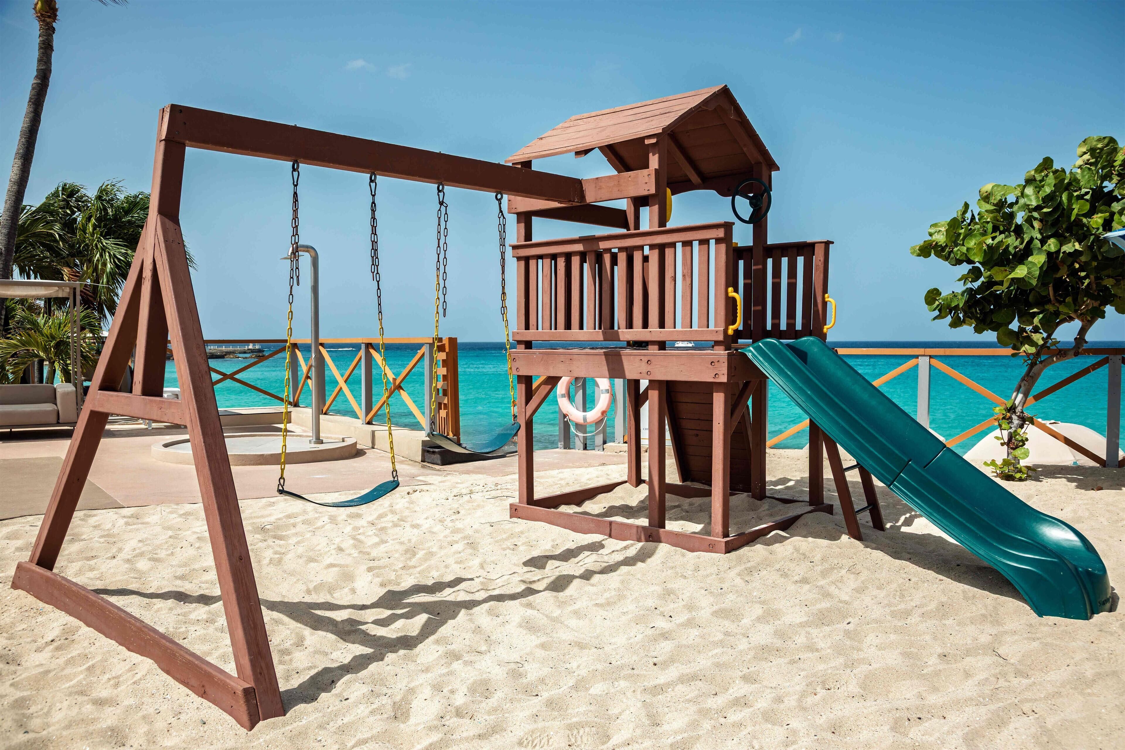 Un ensemble de jeux d’extérieur en bois, avec une balançoire et un toboggan, installé sur une plage de sable, offrant une vue dégagée sur l’océan.