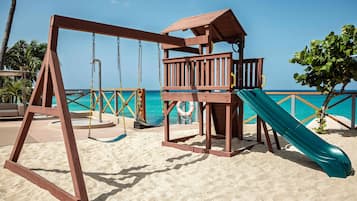 Un ensemble de jeux d’extérieur en bois, avec une balançoire et un toboggan, installé sur une plage de sable, offrant une vue dégagée sur l’océan.