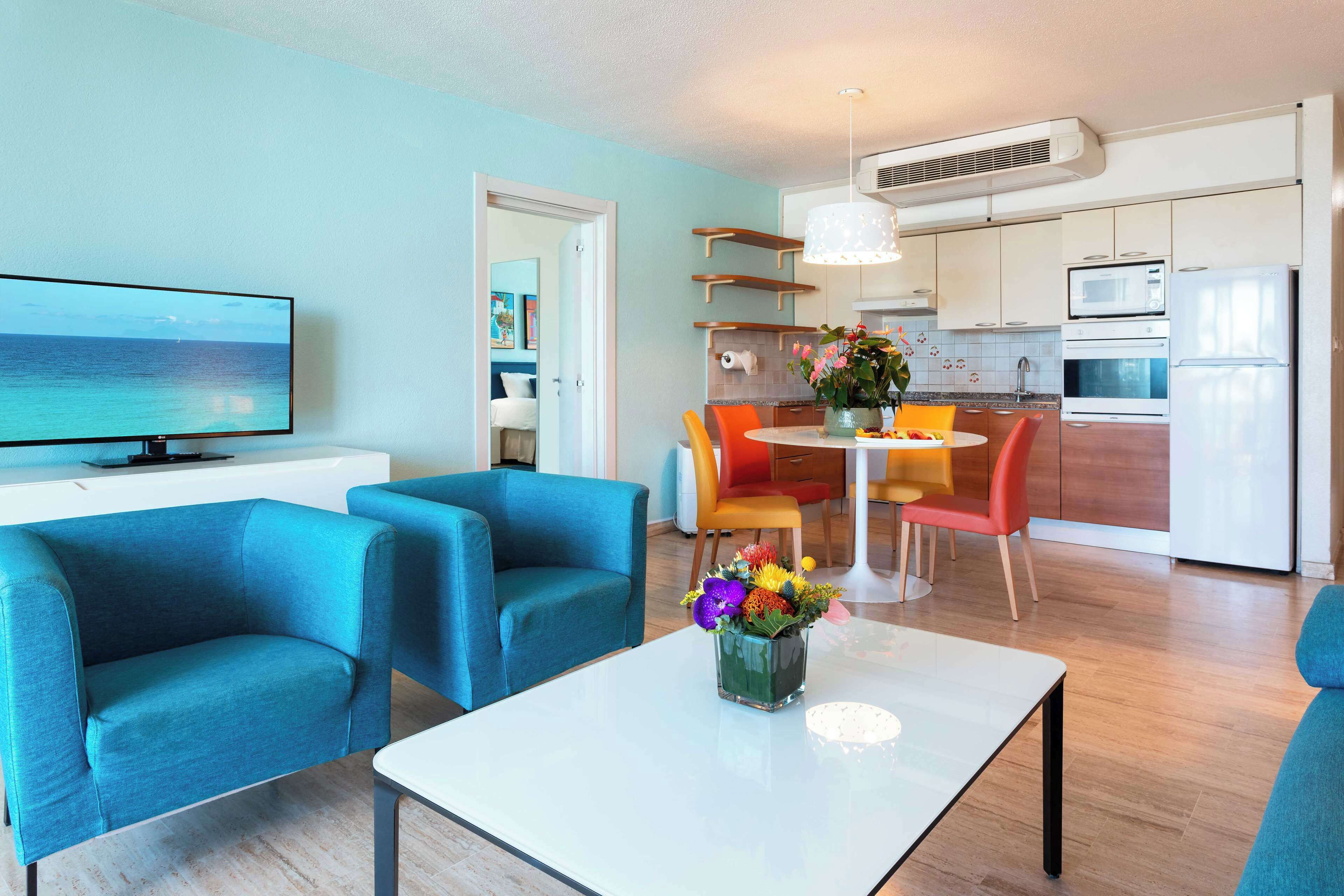 Una sala moderna con un televisor de pantalla plana, sillones azules, una mesa de centro blanca con un florero y un comedor con sillas naranjas.