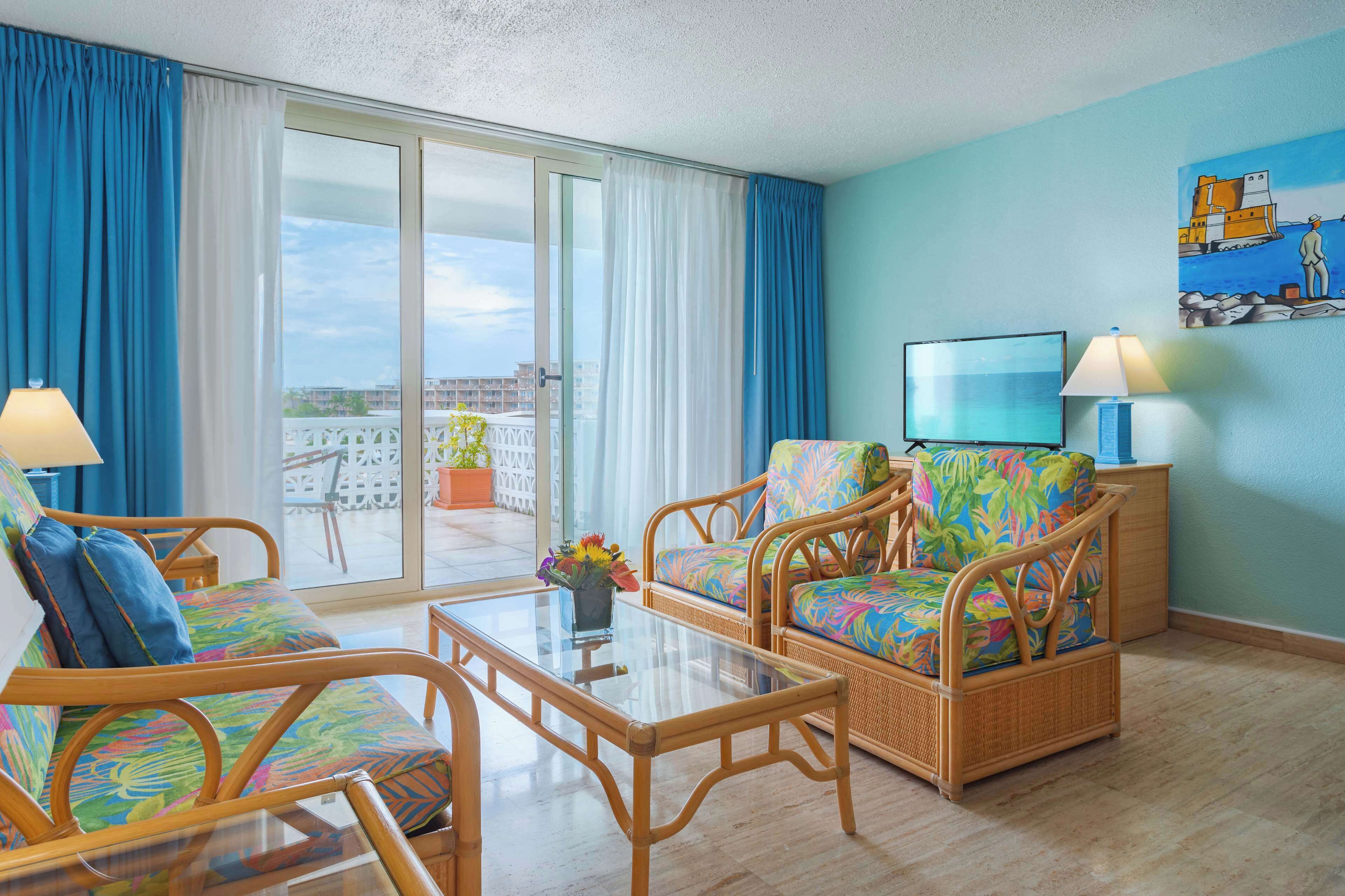 A room with wicker furniture, a glass coffee table, and a view of a cityscape through large windows.