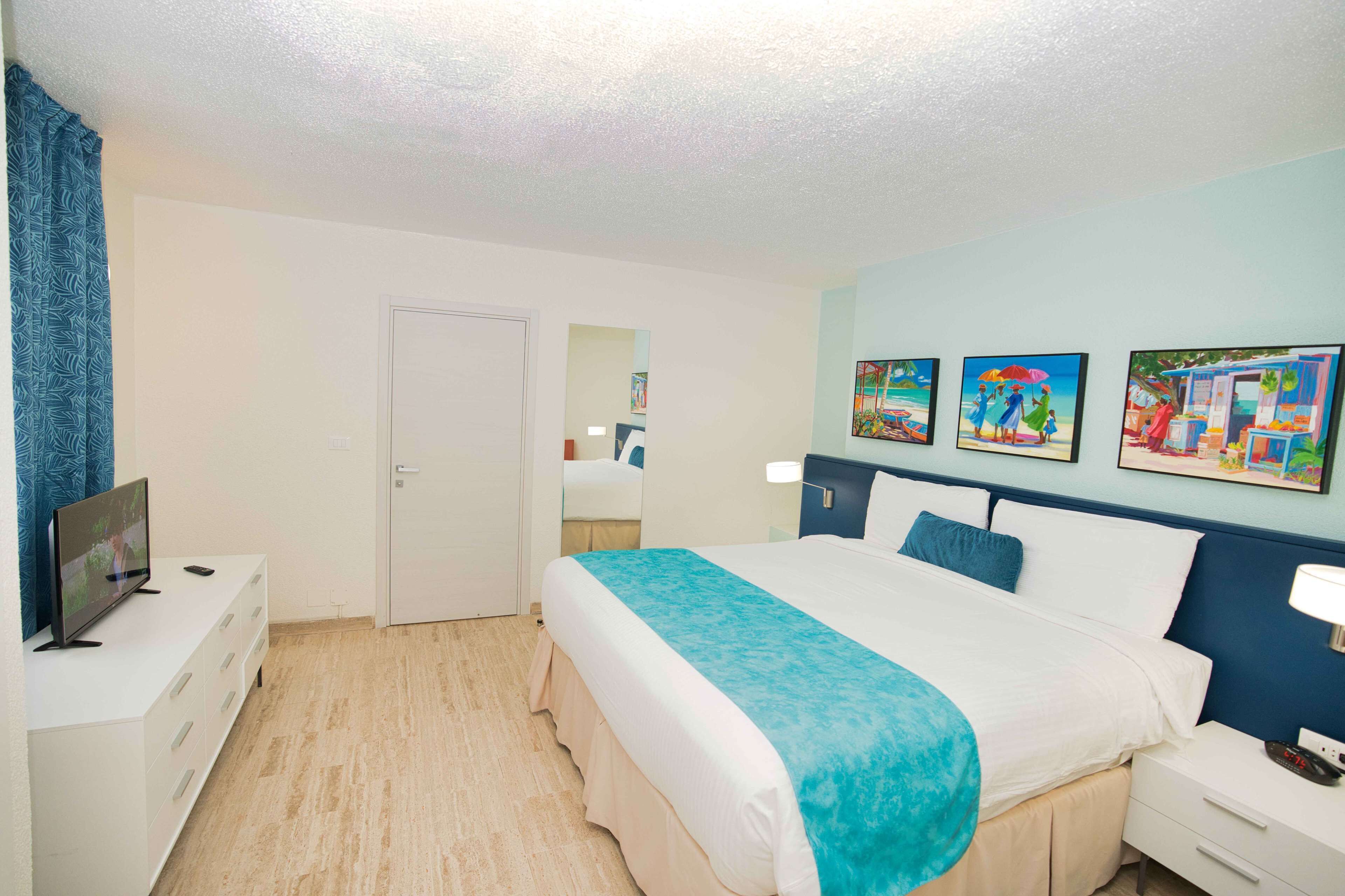 Une chambre d’hôtel avec un lit, un téléviseur posé sur une commode, une porte et trois tableaux accrochés au mur.
