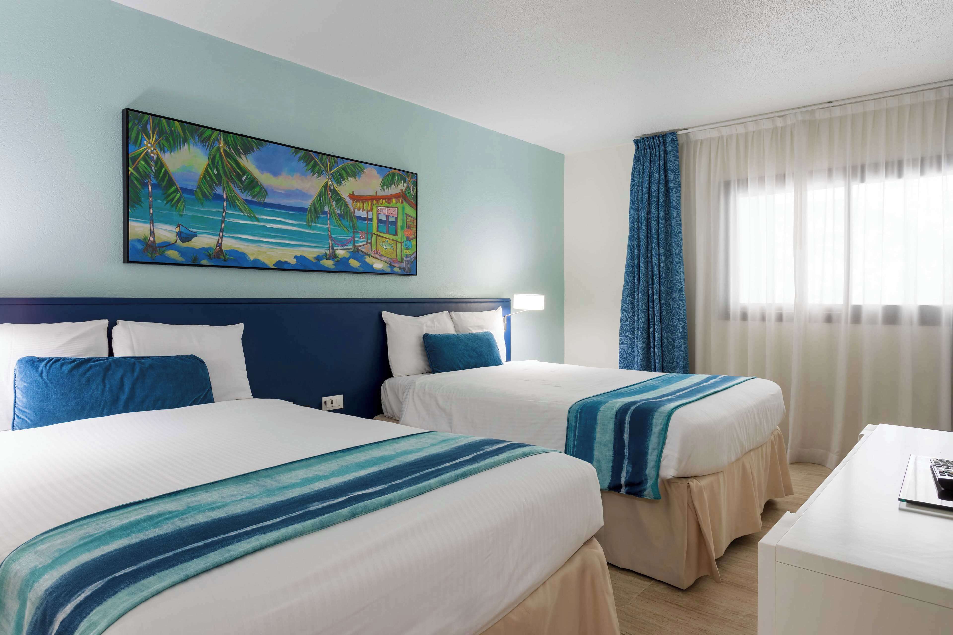 A hotel room with two beds, a painting of a beach scene, and a window with curtains.