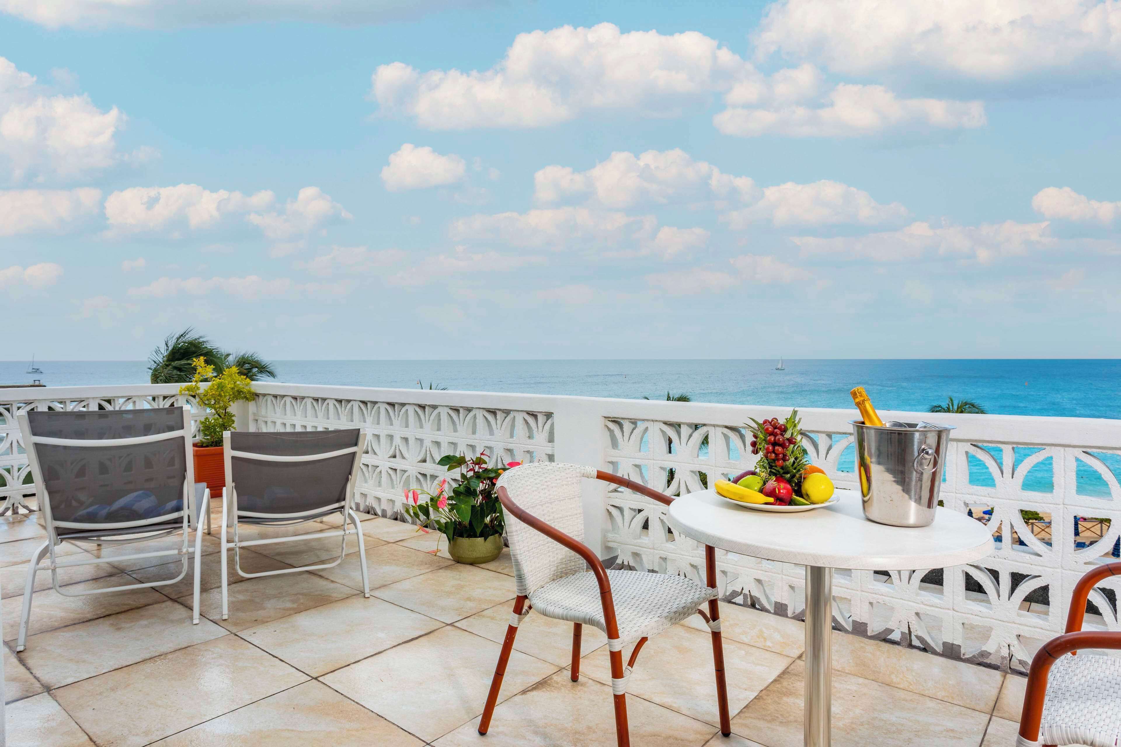 Een terras met twee rieten stoelen, een kleine ronde tafel met een fruitschaal en een champagnefles, en uitzicht op zee.