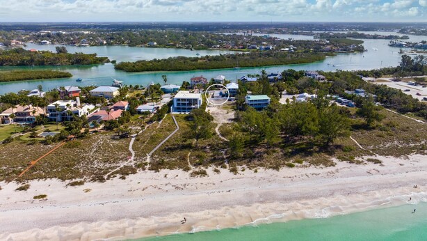 Ett flygbildsbild av en kustort med fastigheter som ligger direkt vid stranden, en småbåtshamn och en fri utsikt över det omgivande vattnet och landskapet.