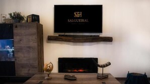 Ein Wohnzimmer mit an der Wand befestigtem Fernseher, darunter ein Kamin und ein Couchtisch aus Holz mit Dekorationselementen.
