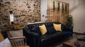 Ein Wohnzimmer mit einer Steinwand, einem schwarzen Ledersofa mit gelben Kissen, einer Holzwand mit Pflanzen und einem Spiegel links.