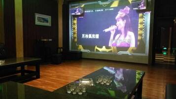 A room with a stage, a screen displaying a performer, and a table with glasses.