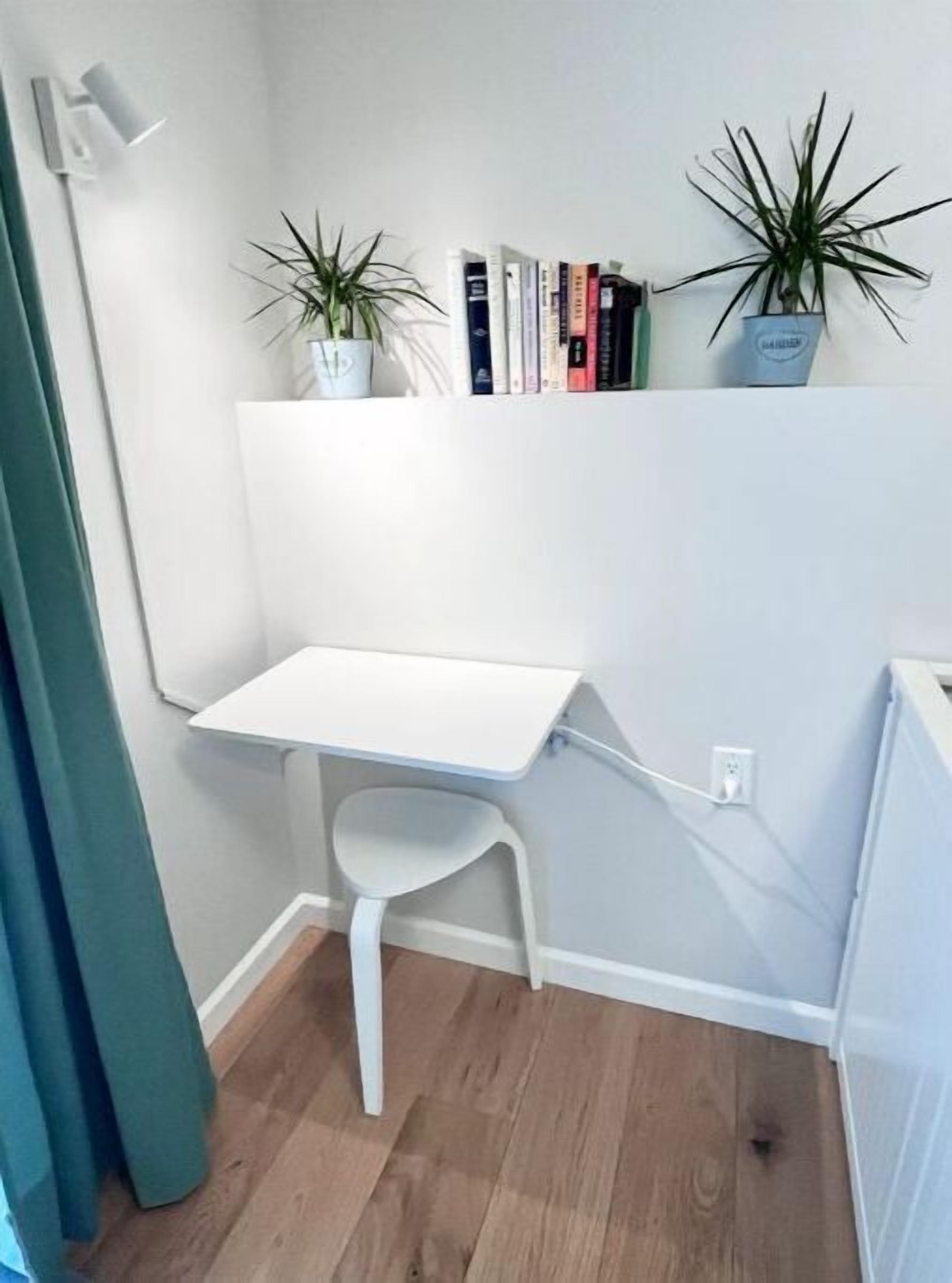 Een klein, wit bureau met een stoel in een hoek van een kamer met houten vloeren. Op een plank boven het bureau staan twee potplanten, en er is een rij boeken op de muur achter het bureau.