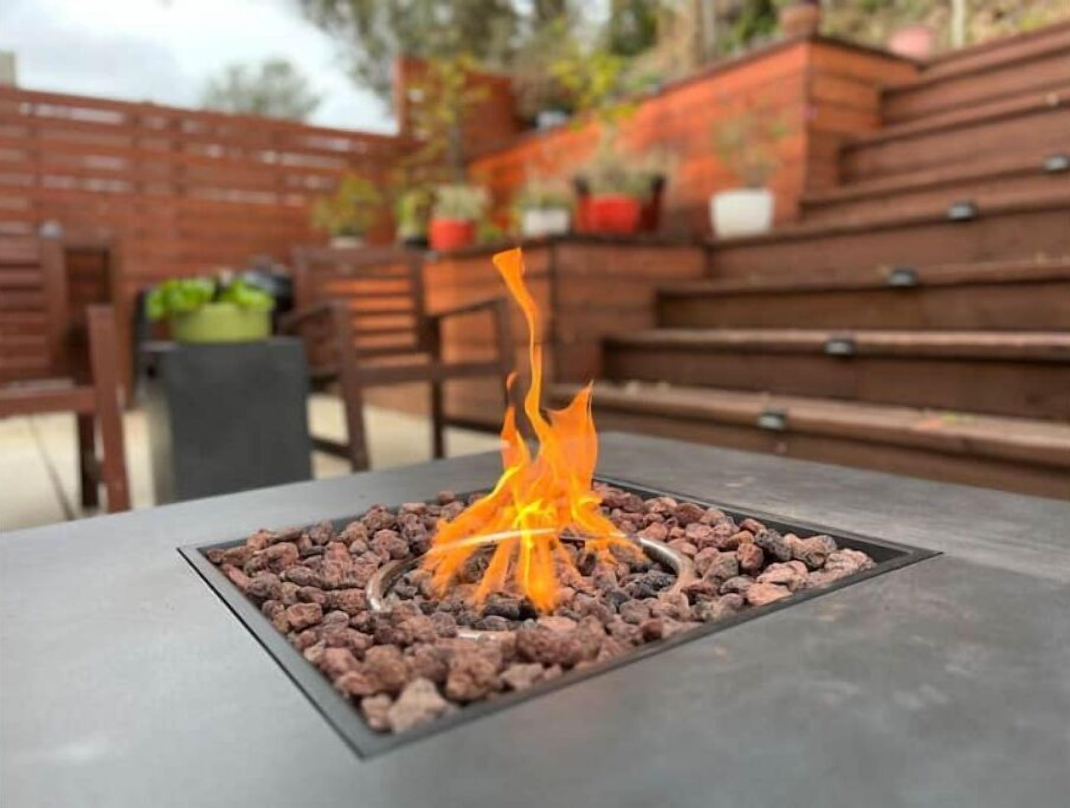 A fire feature with a metal grate and rocks, surrounded by wooden furniture and a brick wall.