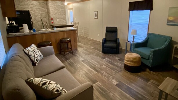 Un salon comprenant un canapé, un fauteuil, un petit pouf, un coin cuisine avec un réfrigérateur et une fenêtre équipée de stores.