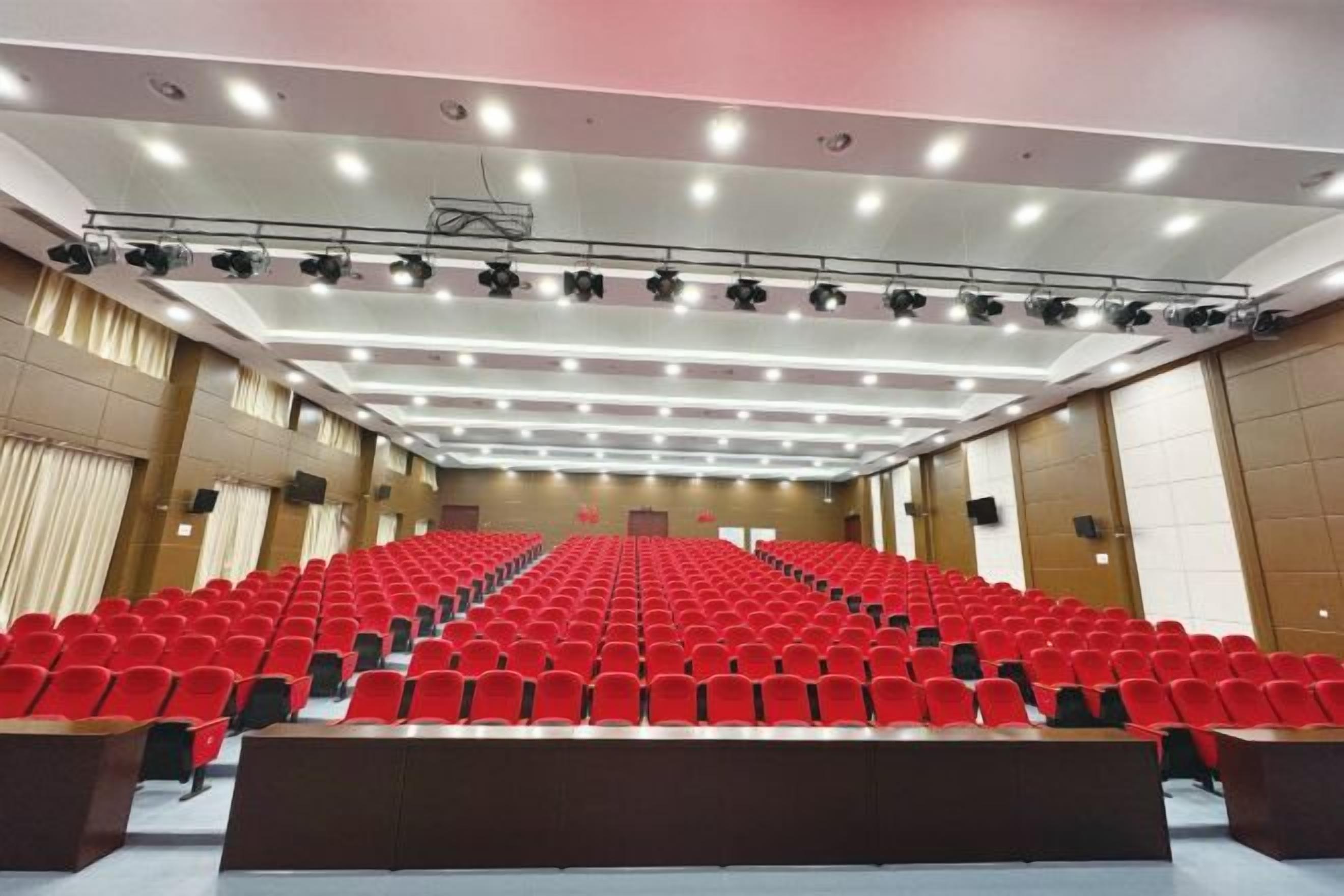 A large auditorium with rows of red chairs, a stage with a podium, and a ceiling with multiple lights.