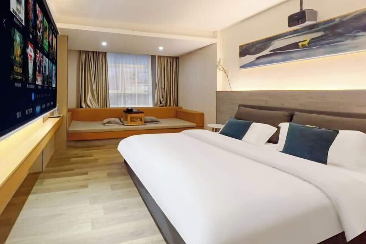A hotel room with a large bed, a seating area with cushions, a TV on the wall, and a window with curtains.