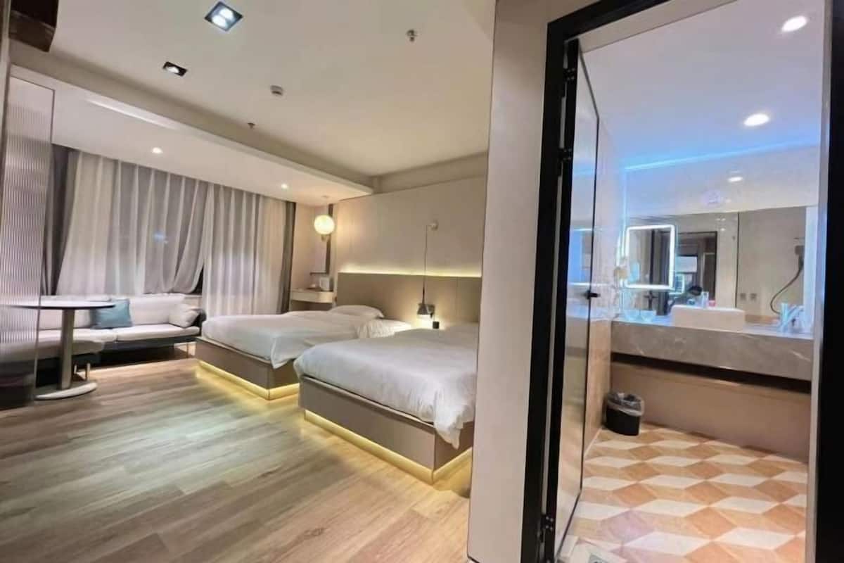 A modern hotel room with two beds, a sofa, a small table, and a bathroom with a mirror.