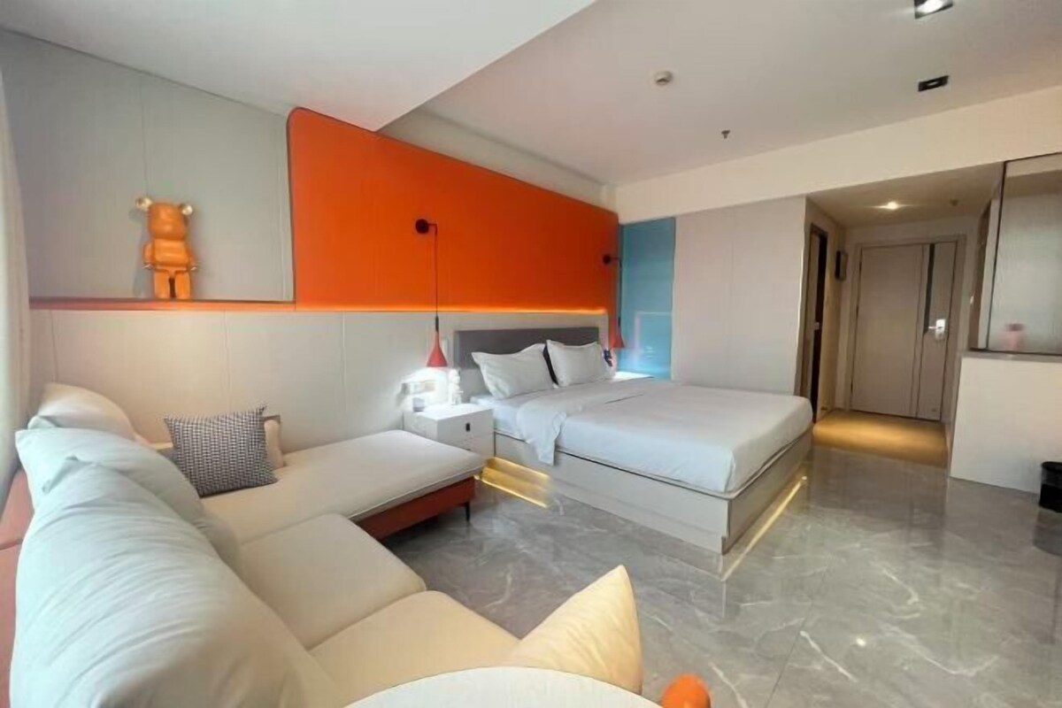 A modern hotel room with a sofa, a bed, a nightstand, and a wall-mounted lamp.