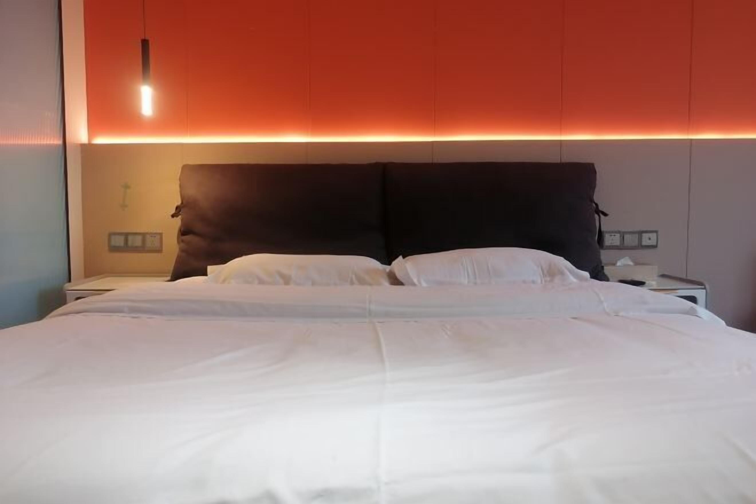Una cama bien hecha con sábanas blancas, cabecera oscura y una lámpara fijada a la pared.