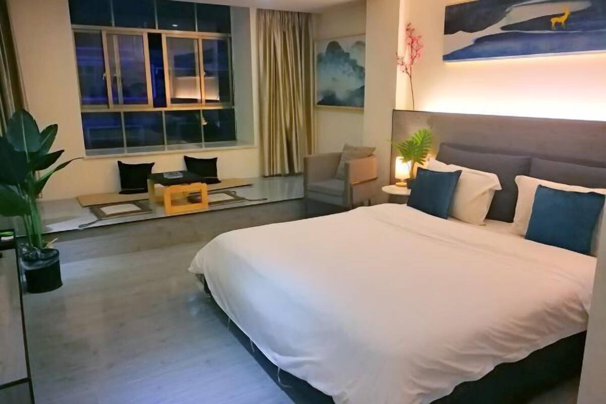 A hotel room with a large bed, a desk with a chair, a window with curtains, and a painting on the wall.