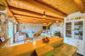 Un intérieur rustique avec un mur en pierre, un plafond en bois, un coin repas avec un banc, une cuisine équipée d’armoires et un escalier.