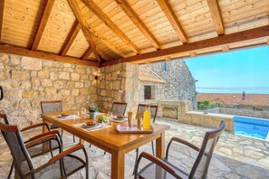 Un espace extérieur aménagé pour les repas, avec des murs en pierre, une table et des chaises en bois, un four en pierre et une vue sur une piscine.