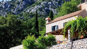 A stone building with a tiled roof, surrounded by lush greenery and a rocky mountain backdrop.