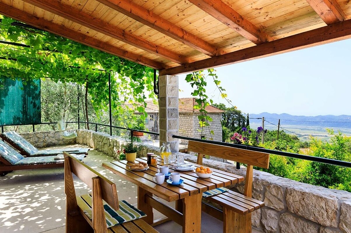 An outdoor patio with a wooden table set for breakfast, stone walls, and a view of the countryside.