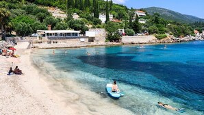A coastal beach with clear blue water, people swimming and paddleboarding, and buildings on a hillside in the background.