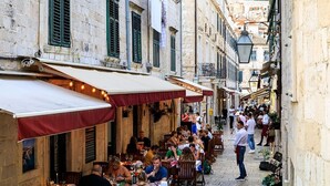 A narrow street lined with outdoor cafes, stone buildings, and a view of a hill in the background.