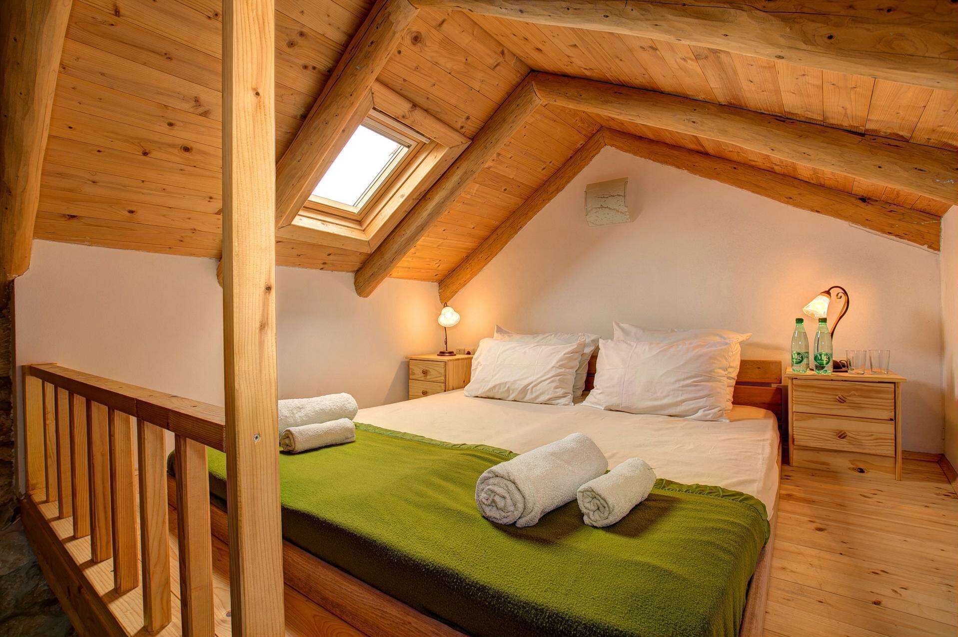 Une chambre confortable située sous les toits, avec un plafond en pente, un puits de lumière, un lit recouvert de draps blancs, deux tables de chevet avec des lampes et une couverture verte sur le lit.
