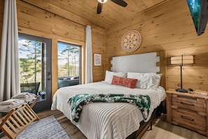 A wooden-paneled bedroom with a bed, bedside table, and a view of trees through a door.