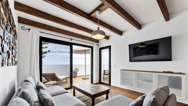 Un salon moderne avec un grand canapé gris, une table basse en bois et un téléviseur fixé au mur. Une porte-fenêtre coulissante donne accès à un espace extérieur aménagé en patio, avec une chaise et une plante.