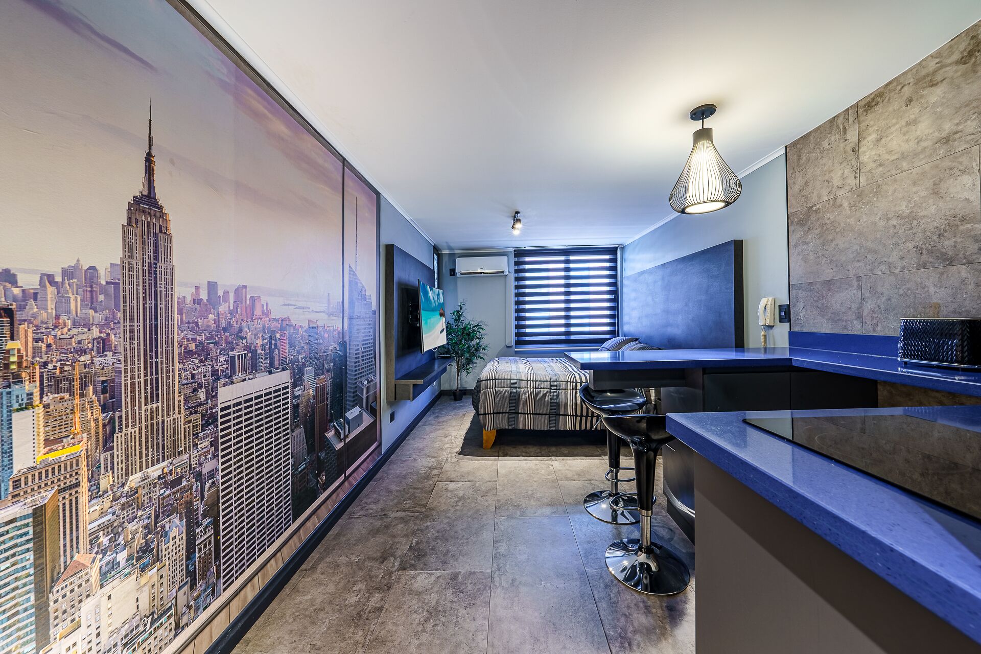 A modern hotel room with a cityscape mural, a large bed, a desk with a chair, and a large window with blinds.