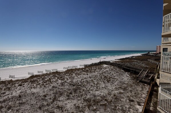 Blick auf einen Strand mit sandigem Ufer, blauem Himmel und einem Gebäude mit Balkonen, das den Strand überblickt.