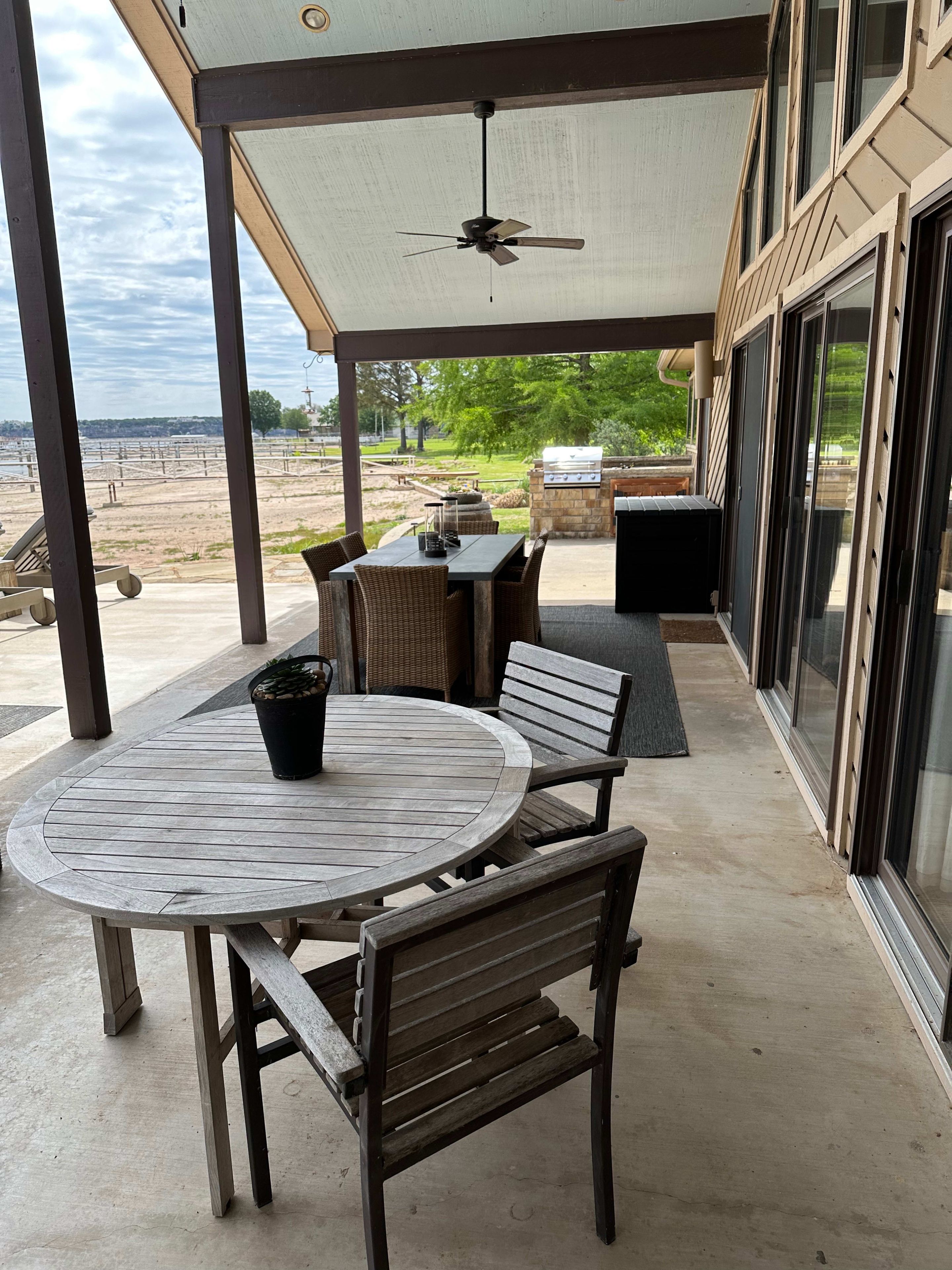 Une terrasse couverte avec une table ronde en bois et des chaises, un ventilateur de plafond, et une vue sur un barbecue et l’espace extérieur.