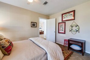 A bedroom with a bed, a small desk, and a chair. There are framed pictures on the wall and a welcome sign.
