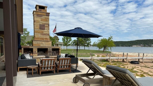 Une terrasse extérieure dotée d’une cheminée en pierre, de fauteuils confortables et offrant une vue sur un lac.