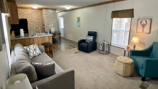 A living room with a sofa, a chair, a ottoman, a lamp, a TV, a kitchen area with a counter and chairs, and a window with curtains. - Rainbow River Bungalow F (Dunnellon)