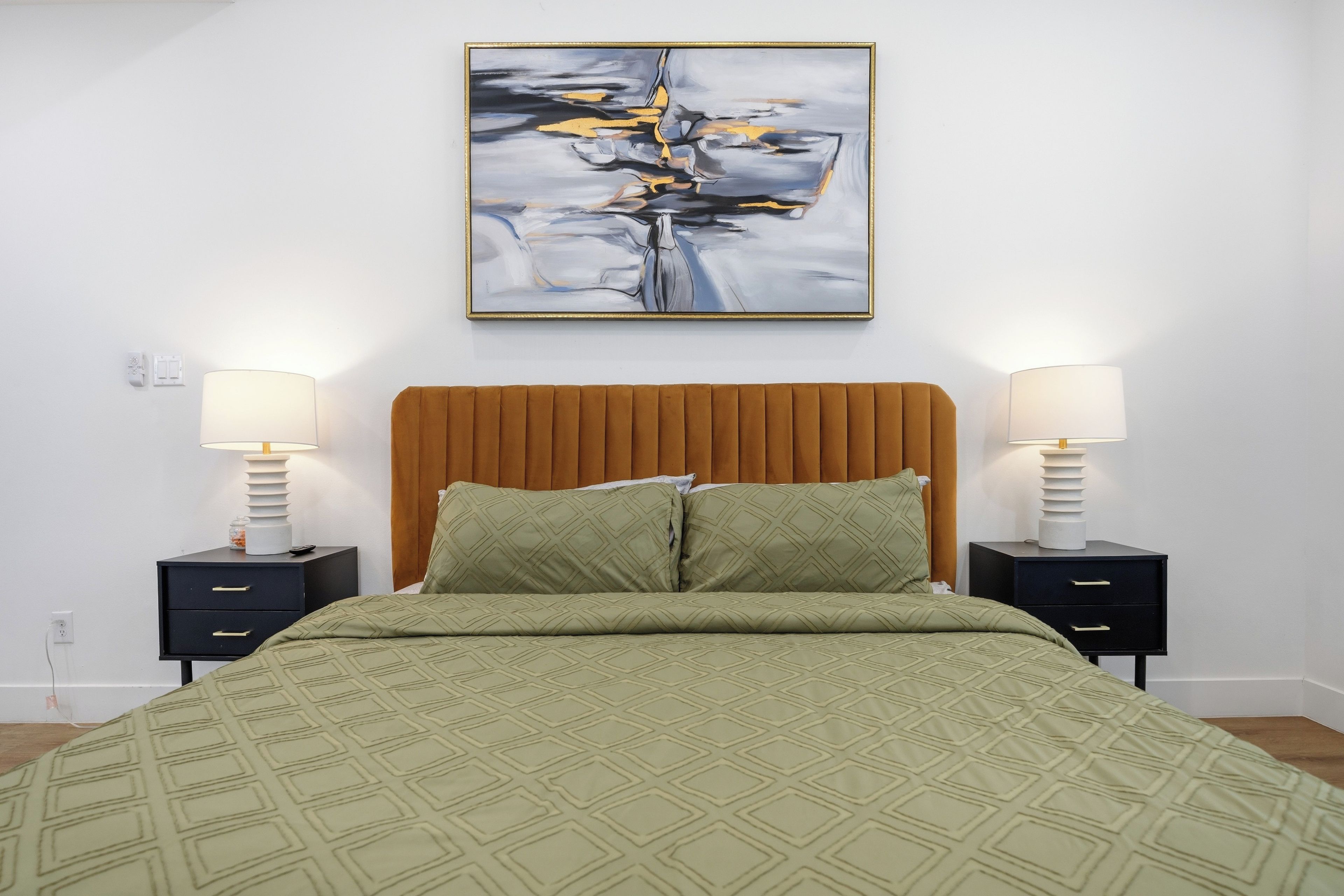 Un letto rifatto con cura, con una coperta a motivi verdi, affiancato da due comodini con lampade, e un quadro astratto moderno sopra il letto.