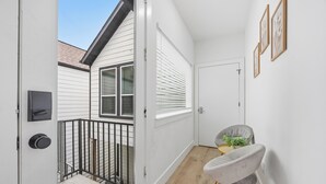A narrow hallway with a door leading to a balcony, a small seating area with a chair and a plant, and a white door with a window.