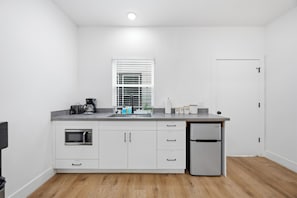 Una cocina compacta con armarios blancos, microondas integrado, cafetera, refrigerador y ventana con persianas.