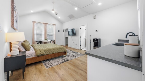 A modern, well-lit bedroom with a bed, bedside table, and a TV. Adjacent is a kitchenette with a sink and countertop.