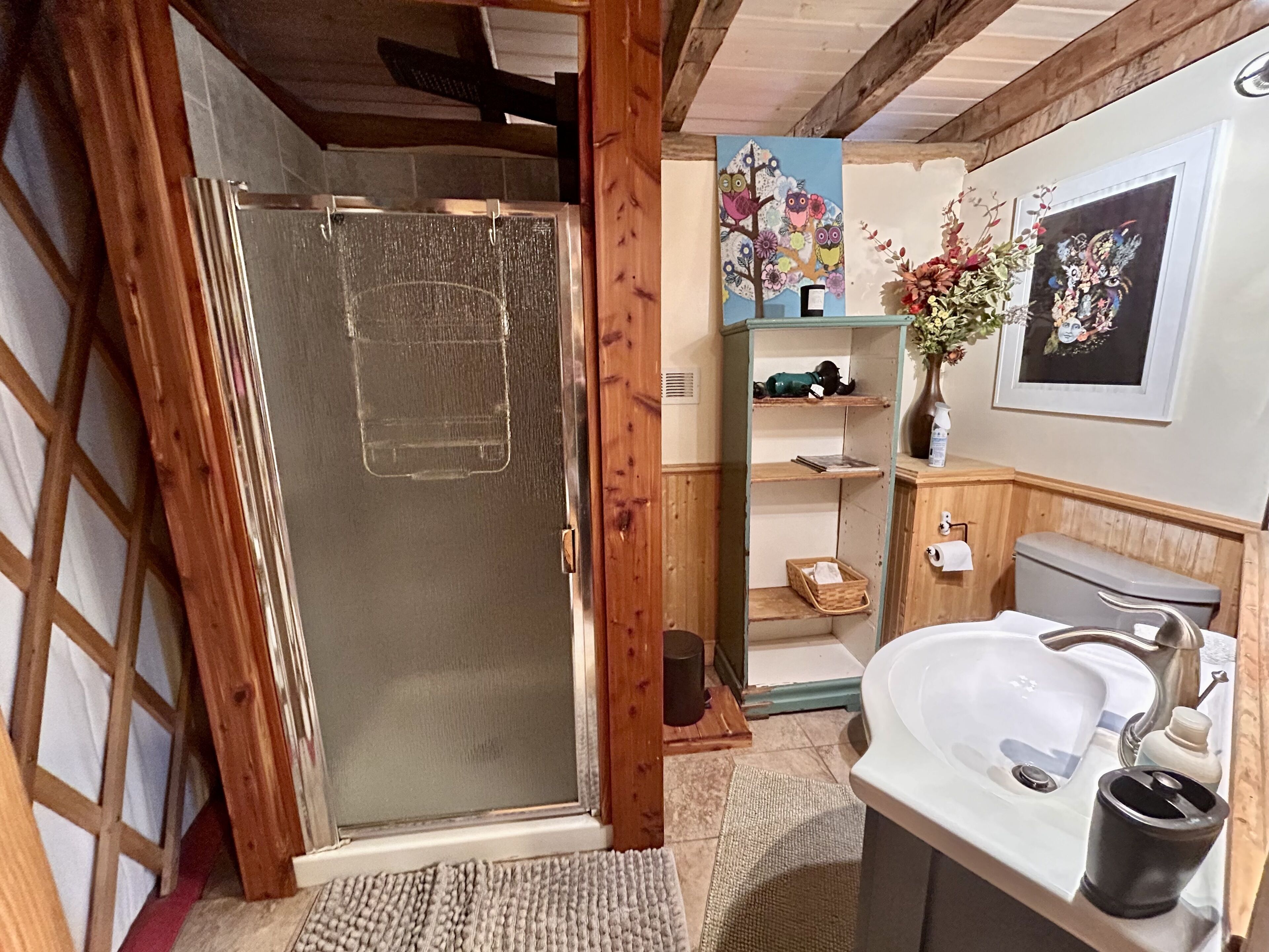 A bathroom with a glass shower enclosure, wooden cabinets, a sink, and shelves with toiletries.