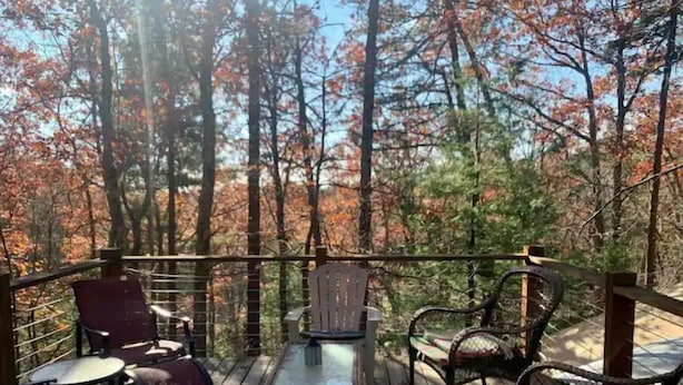 An outdoor deck with wooden furniture, including chairs and a table, surrounded by trees with autumn foliage. - Three-story Blue Ridge Yurt getaway (Pilot)