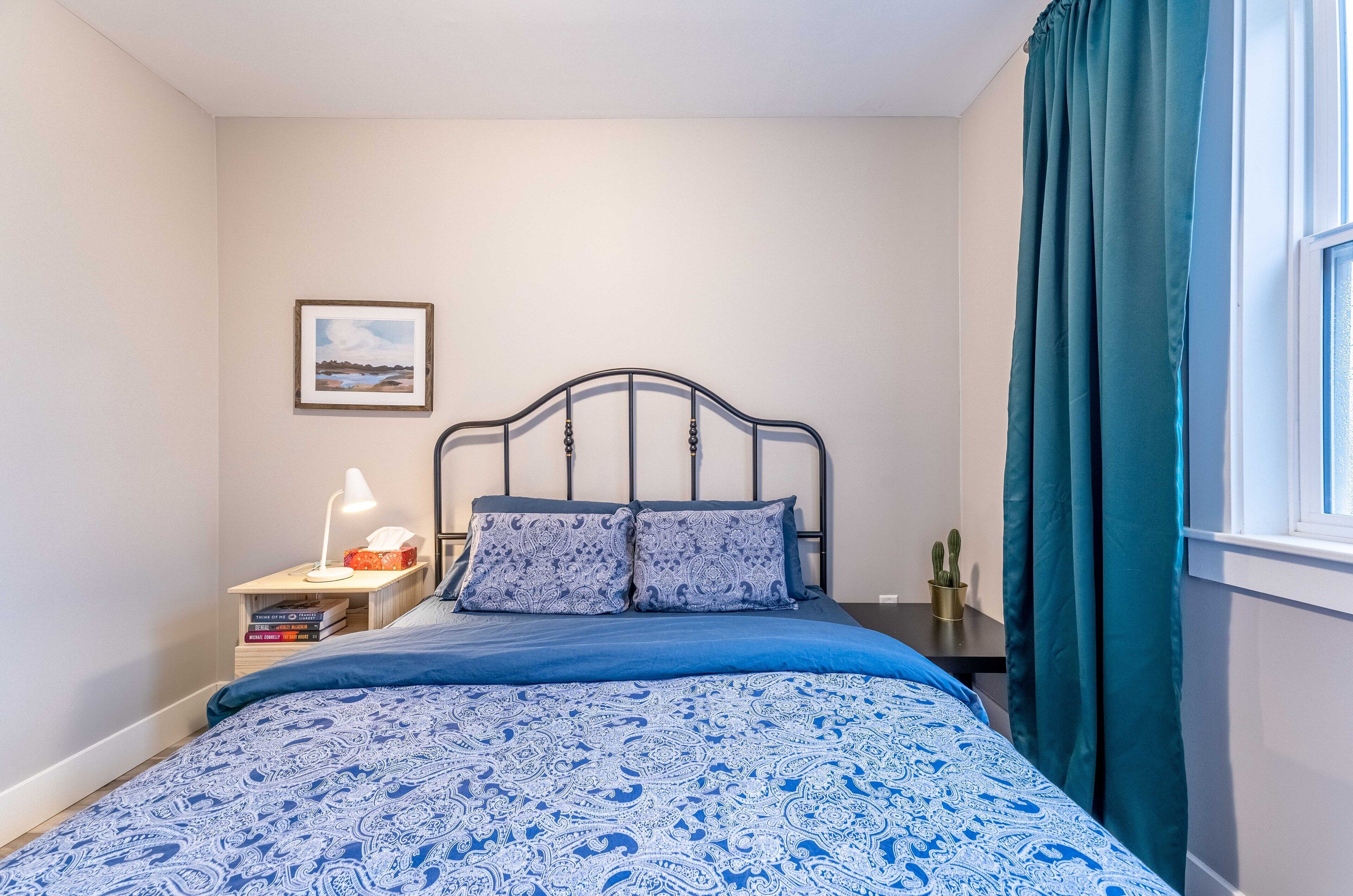 Ein ordentlich bezogenes Bett mit einer blau gemusterten Tagesdecke, ein Nachttisch mit einer Lampe und Büchern sowie ein Fenster mit taubenfarbenen Vorhängen.