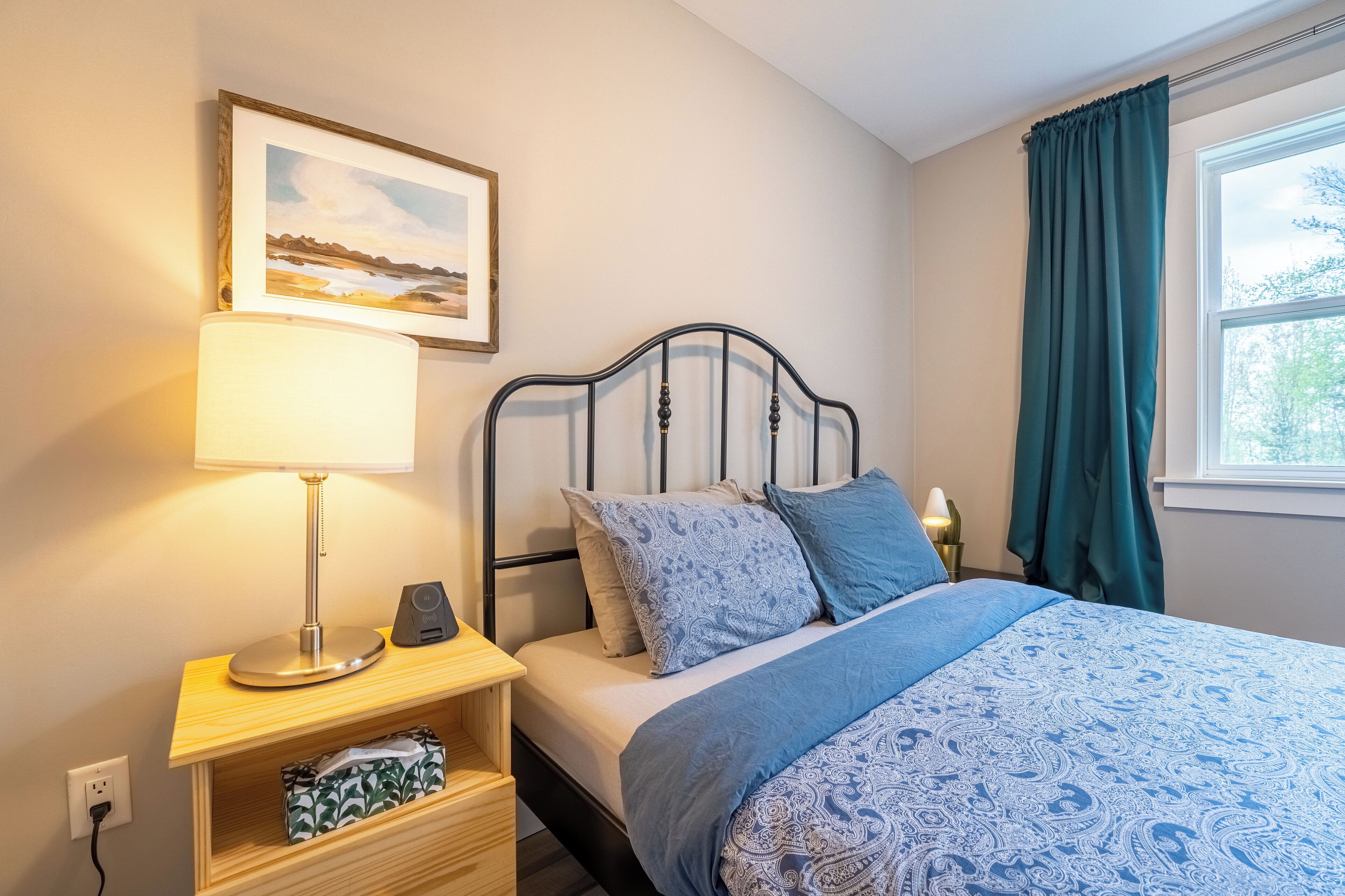 A neatly arranged bedroom with a bed, a bedside table with a lamp, a framed picture on the wall, and a window with curtains.