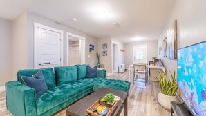 A living room with a teal sectional sofa, a coffee table with a tray, a TV, and a dining area with a table and chairs. - YHZ Cheerful Private Unit 10 min to Airport (ELMSDALE)